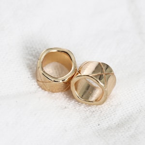 May include: Two gold-coloured, open-ended rings with a textured, quilted design. The rings are made of a shiny metal and are set against a white background. The rings are likely jewellery.