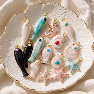 Könnte beinhalten: Eine Sammlung von Metallanhängern in Form verschiedener Meerestiere wie Fische, Seesterne und Muscheln. Die Anhänger sind mit Emaille in Weiß, Schwarz, Rosa und Türkis verziert und mit kleinen eingebetteten Perlen, Türkisen und Kristallen besetzt.
