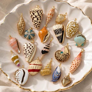 Puede incluir: Una colección de dijes de metal con forma de conchas marinas que presentan diversos patrones de esmalte, colores y texturas. Cada dije incluye una pequeña anilla dorada en la parte superior para colgar, mostrando formas variadas de conchas.