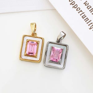Puede incluir: Dos colgantes cuadrados con piedras rosas. Uno tiene un marco dorado con borde blanco y borde texturizado. El otro tiene un marco plateado con borde blanco y borde texturizado. Ambos tienen una piedra rosa rectangular en el centro.