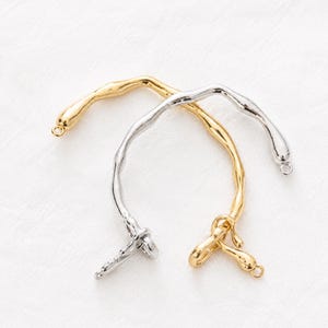 Könnte beinhalten: Zwei gebogene Armbandkomponenten aus Metall mit einer organischen, geschmolzen wirkenden Textur. Ein Teil ist in poliertem Goldton gehalten, das andere in poliertem Silberton. Jedes Ende der gebogenen Stäbe ist mit einer kleinen runden Öse versehen.