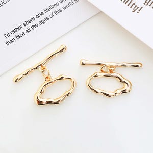 May include: Two gold-colored toggle clasps. Each clasp features a bar and an irregular oval shape. The clasps are on a white surface, with text from a document in the background.
