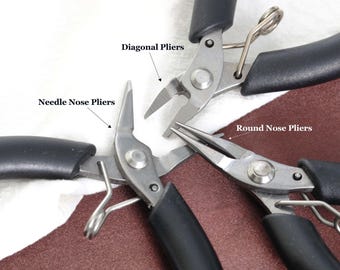 May include: Close-up of three different types of pliers: needle nose, diagonal, and round nose. The pliers have silver metal heads and black handles. The tools are arranged on a white and brown surface.