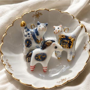 Pode incluir: Três pequenos pingentes de cerâmica em forma de animal em uma tigela branca festonada com borda dourada. Dois pingentes são gatos com detalhes pintados em azul e ouro, e um é um cavalo com um design de lua azul, crina dourada e pernas listradas de vermelho e branco.