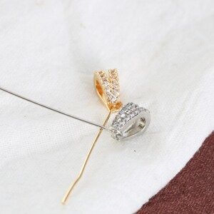 Peut inclure: Gros plan sur deux apprêts de bijoux. L'un est doré avec des pierres transparentes, l'autre est argenté avec des pierres transparentes. Une longue et fine épingle argentée est également présente, posée sur un tissu blanc.