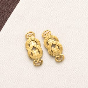 May include: Two identical gold-toned metal fasteners featuring an intricate, symmetrical knot-like design with looped ends and vertical parallel lines. These decorative hardware pieces have a polished finish and a distinct, elongated oval shape.