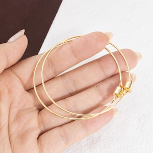 May include: A pair of thin, circular gold-toned wire bracelets. Each bracelet features a small, cylindrical magnetic clasp mechanism that connects the two ends of the wire, creating a continuous, smooth, and minimalist hoop design.