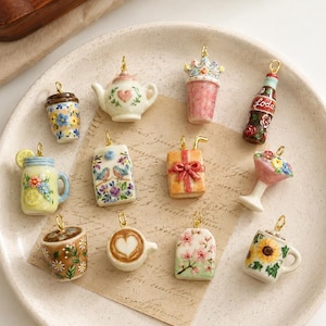 May include: A collection of twelve miniature ceramic charms featuring various designs including a teapot, a soda bottle, a gift box, a mason jar, a milk carton, a coffee cup with latte art, and mugs decorated with floral patterns and sunflowers.
