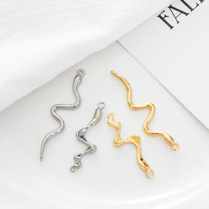 Puede incluir: Cuatro colgantes metálicos con forma de serpiente en tonos plateados y dorados. Los colgantes tienen un diseño ondulado y un pequeño lazo en la parte superior para sujetar. Fondo blanco.