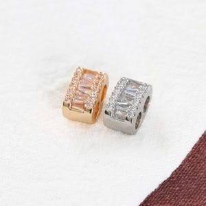 May include: Two rectangular jewelry beads, one gold-toned and one silver-toned, each adorned with clear, rectangular and round-cut crystals. The gold bead has a warm, champagne hue, while the silver bead sparkles. These beads are likely for jewelry making.