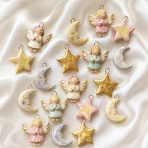 May include: A collection of small ceramic charms featuring hand-painted angels with wings, crescent moons with closed eyes, and five-pointed stars. The charms are finished in pastel pink, blue, yellow, and metallic gold, each with a silver metal jump ring.