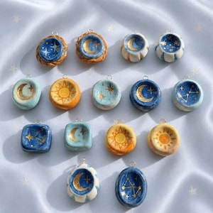 Könnte beinhalten: Eine Sammlung von fünfzehn kleinen, handgefertigten Keramikanhängern mit Himmelsmotiven. Jedes Stück zeigt handbemalte goldene Monde, Sonnen und Sterne auf blauem, weißem und orangefarbenem Hintergrund, versehen mit einer Metallöse zum Aufhängen.