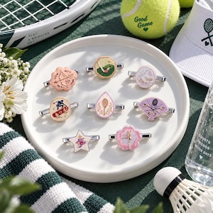 Peut inclure: Huit broches en émail métallique aux formes et motifs variés, incluant un arc et une flèche, un club de golf, des patins à glace, un skateboarder, une raquette de tennis, un éventail, une étoile et une ballerine, sur une assiette ronde.