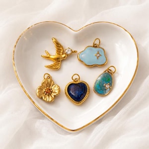 May include: Five gold-toned metal charms resting in a white heart-shaped dish. The charms include a bird with a clear stone, a light blue cloud with a star, a gold flower with an orange center, a dark blue heart, and a blue-green marbled oval.