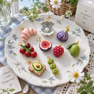 May include: A collection of miniature food-themed charms including a pink croissant, an open oyster shell with a pearl, a purple fig, a cluster of red tomatoes, a sliced fig, a red raspberry, a green olive, avocado toast, and three green peas.