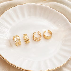 May include: A set of four gold-toned ear cuffs. The first is a double band, the second features a leaf-like pattern, the third has a cut-out star design, and the fourth is a plain, smooth band. Each piece is open-ended for adjustable placement.