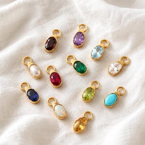 May include: A collection of twelve oval-shaped jewelry charms, each featuring a gold-toned metal setting with a circular jump ring at the top. The charms contain various colorful stones, including deep red, purple, light blue, green, white, and yellow.