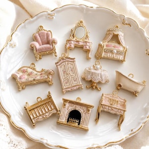 Puede incluir: Un conjunto de diez dijes de muebles en miniatura hechos de metal dorado con detalles en esmalte rosa y crema. El set incluye un sillón con textura de terciopelo, un tocador con espejo, una cama con dosel, una chaise longue floral, un armario, una mesa, una bañera, una cuna, una chimenea y un escritorio.
