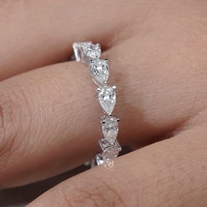 Könnte beinhalten: Ein silberfarbener Eternity-Ring mit einer Reihe von klaren, birnenförmig geschliffenen Steinen in einer durchgehenden Fassung. Der Ring wird an einem Finger getragen und zeigt das facettierte Design der Steine sowie das Metallband der Fassung.