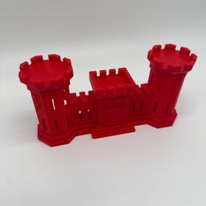 May include: A bright red, three-dimensional castle structure featuring two tall cylindrical towers on the ends and a central gatehouse. The design includes crenelated battlements along the top edges and a small arched doorway in the center.