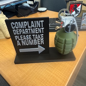May include: A novelty desk sign that reads "COMPLAINT DEPARTMENT PLEASE TAKE A NUMBER" in silver lettering on a black background. A green grenade with a silver handle and the number "1" is next to the sign.