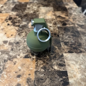 Puede incluir: Una granada verde oliva con un anillo de tracción plateado y una parte superior verde. La granada está sobre una superficie estampada marrón y beige.