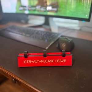 May include: A red desk sign with the text "CTR+ALT+PLEASE LEAVE" in white. The sign has three black clips on top. The sign is on a desk with a keyboard, mouse, and computer monitor in the background.