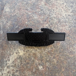 May include: A black, 3D-printed badge featuring a keyboard design at its centre, flanked by rectangular panels. The badge has a textured surface and is set against a mottled, grey-brown background.