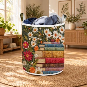 May include: A cylindrical fabric laundry hamper featuring a quilted exterior with a printed pattern of white daisies, orange flowers, and red blossoms surrounding a stack of colourful vintage books. The top is open, revealing folded clothing.