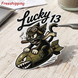 May include: A die-cut sticker featuring a black cat wearing aviator goggles and a scarf, smoking a cigar while riding an olive green bomb with a shark mouth graphic. The text Lucky 13 is printed in black script above the cat on a white background.