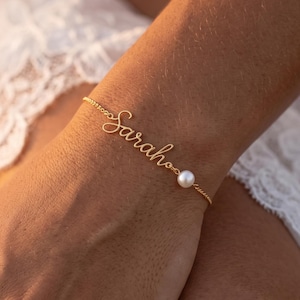 May include: A gold-toned chain bracelet featuring a delicate script nameplate that reads Sarah, accented with a single small white pearl bead integrated into the chain link design, creating a refined and simple piece of wrist jewelry.