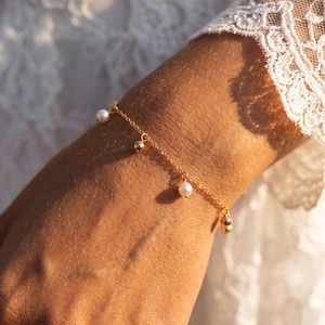 Könnte beinhalten: Ein zartes Goldkettenarmband mit kleinen, hängenden weißen Perlen und polierten Goldkügelchen. Das Armband besteht aus einer feingliedrigen Kette, die um das Handgelenk liegt und eine Anordnung aus abwechselnden Perlen- und Goldkugelanhängern aufweist.