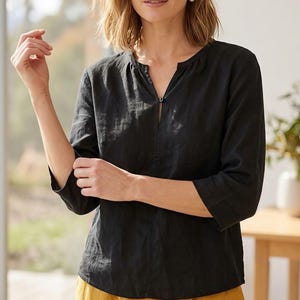 May include: Black linen top with a split neckline and three-quarter sleeves. The top has a relaxed fit and is paired with yellow pants. The top features a small button closure at the neckline. The fabric appears to be lightweight and breathable.