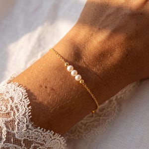 May include: A delicate gold chain bracelet featuring three small, round white pearls centered between two tiny, textured gold beads. The bracelet is constructed with a fine link chain that wraps around the wrist, showcasing a minimalist design.