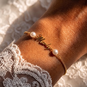 May include: A gold-toned chain bracelet featuring a central nameplate with the script text Marly, flanked by two white spherical pearls. The delicate metal chain connects to the pearls and the central name element, creating a refined accessory.