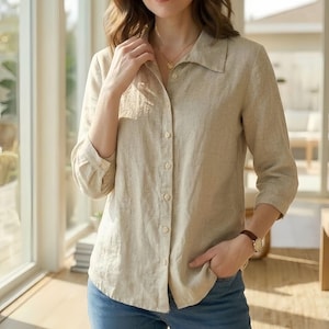 Könnte beinhalten: Beige Leinenhemd mit Kragen und Dreiviertelärmeln. Das Hemd ist zugeknöpft und wird mit einer blauen Jeans getragen. Eine goldene Halskette und eine braune Uhr sind ebenfalls zu sehen.