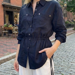 Può includere: Una camicia a maniche lunghe blu navy scuro con abbottonatura, colletto, due tasche sul petto e un cordoncino in vita. La camicia è abbinata a pantaloni bianchi dal taglio ampio, creando un insieme pulito e funzionale.