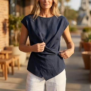 May include: A navy blue linen top with a unique wrap design. The top features short sleeves and a round neckline. The model is wearing white pants. The top is made of a soft, lightweight fabric, perfect for warm weather.