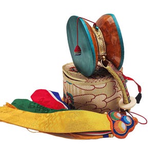 Puede incluir: Un tambor damaru tibetano tradicional con parches turquesa y marrón, detalles dorados y un mango decorativo. Descansa sobre una caja cilíndrica estampada, acompañado de cintas y borlas de seda coloridas. Un artefacto cultural.