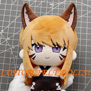 May include: A plush doll with light brown hair, pointed fox-like ears, and purple eyes. The doll wears a white top with a black collar and dark brown trousers featuring white floral embroidery. The text CUSTOM FELT DOLLS! is printed in orange.