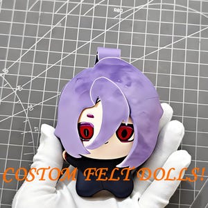 May include: A felt doll with a purple hairstyle and red eyes. The doll is black and white with the text "CUSTOM FELT DOLLS!" in orange. The doll is held in a gloved hand.
