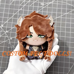 May include: A small plush doll with brown hair, pointed ears, and green eyes, wearing a dark patterned outfit. The doll features braided hair sections and a smiling expression. The text "COSTOM FELT DOLLS!" is printed in orange across the front.