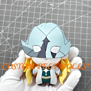 May include: A custom felt doll with a light blue helmet, blonde hair, and a white and teal outfit. The doll has the text "CUSTOM FELT DOLLS!" in orange. The doll is held in a gloved hand.
