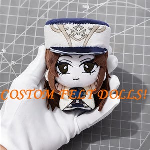 May include: A custom felt doll with a white and navy blue hat, brown hair, and large eyes. The doll is held in a white gloved hand. The text "CUSTOM FELT DOLLS!" is printed in orange.