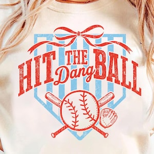 May include: Cream-coloured t-shirt with a baseball-themed graphic. The design features the text "Hit the Dang Ball" in red, a baseball, bats, and a glove. The graphic is set against a light blue background.