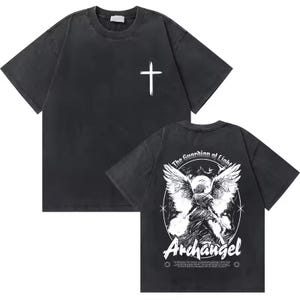 Op de afbeelding: Donkergrijs t-shirt met een witte kruis op de voorkant. De achterkant heeft een gedetailleerde afbeelding van een engel met de tekst "The Guardian of Light" en "Archangel". Het shirt heeft een vintage, versleten uitstraling.