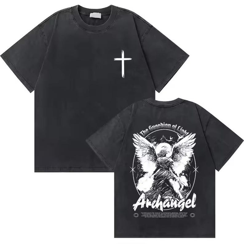 Op de afbeelding: Donkergrijs t-shirt met een witte kruis op de voorkant. De achterkant heeft een gedetailleerde afbeelding van een engel met de tekst "The Guardian of Light" en "Archangel". Het shirt heeft een vintage, versleten uitstraling.