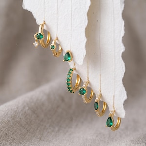 May include: A collection of seven gold hoop earrings suspended from a torn white paper edge. The earrings feature various designs, including small green circular stones, clear crystals, and pear-shaped green gems set in polished gold metal hoops.