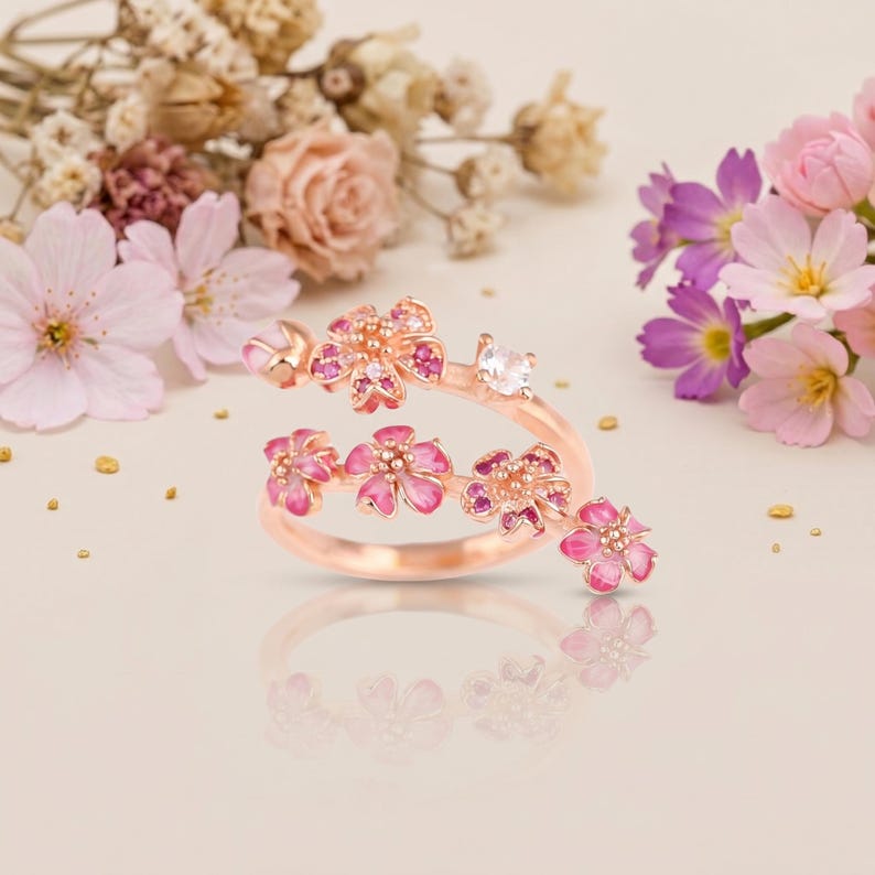 K&ouml;nnte beinhalten: Ein ros&eacute;goldener Ring mit floralem Design, der rosa emaillierte Bl&uuml;tenbl&auml;tter und kleine, funkelnde Edelsteine aufweist. Der Ring umschlie&szlig;t den Finger und erzeugt einen zarten und femininen Look. Der Ring ist vor einem weichen, neutralen Hintergrund.