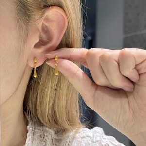 Può includere: Orecchini a goccia color oro. Gli orecchini hanno un piccolo perno rotondo nella parte superiore e un pendente a forma di goccia. Gli orecchini vengono tenuti vicino all'orecchio.